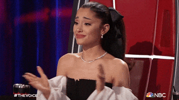 Reality TV gif. Ariana Grande as a judge on The Voice smiles and applauds teary-eyed as she looks at someone off screen. 