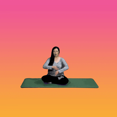 Digital compilation gif. A real woman sits on a yoga mat with eyes closed, raising her hands and drawing them into a prayer position as illustrated flowers and text emerge from her yoga mat against a yellow-pink gradient background. Text, "Good morning. Take it easy on yourself."