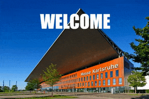 Photo gif. Above the front facade of a building labeled “Messe Karlsruhe” reads the message, “Welcome Back!”