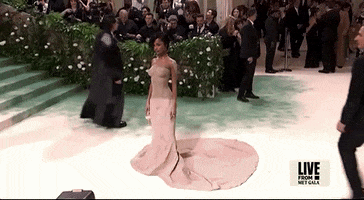 Met Gala 2024 gif. Camera zooms in on Tyla wearing a sand-colored sculpted Balmain mermaid-style dress with shimmery sand makeup on her shoulder. The dress is fitted all the way through her ankles and then pours out into a circular train behind her. 