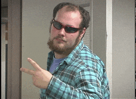 Video gif. A man in wraparound sunglasses holds up a peace sign with two fingers, then says, "Peace," before turning to walk away.