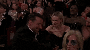 Oscars 2024 GIF. Carey Mulligan, smiling, leans in to pat Bradley Cooper on the back with sisterly pride, as he leans back over the seat to celebrate with her, blushing with bashful happiness.