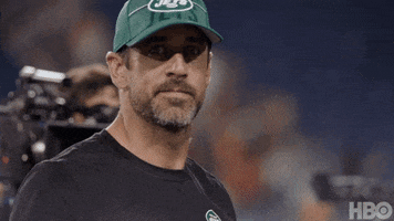 Sports gif. Coach Aaron Rodgers in a Jets baseball cap walking and giving us a wave with a friendly expression as cameras follow behind.
