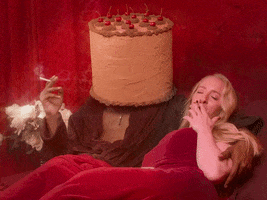 Video gif. Woman smokes a cigarette in bed with a muscular man in a rope with the bed of a Chocolate cake with cherries on top. The sexy cake man holds a cigarette in his hand too and nods his chocolate cake head in a satisfied manner. The woman blows out smoke from her mouth slowly and with a satisfied smile.