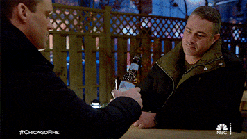 TV gif. Taylor Kinney as Kelly and Jesse Spencer as Matthew in Chicago Fire hold beers as they bring them together in a toast. 