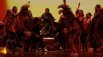 Oscars 2024 gif. Scott George and Osage singers perform 'Wahzhazhe (A Song For My People)' from Killers of the Flower Moon. While an inner circle of men play a powwow drum, a group of singers in Indigenous wear sing and perform choreographed footwork around them. 