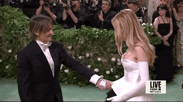 Met Gala 2024 gif. Keith Urban grabs a shy Nicole Kidman's hand, who, now with bolstered confidence, tosses her hair, revealing her face.