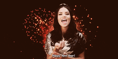 Video gif. As fireworks go off in the background, a woman in a sequined shirt holds up a handful of confetti and blows it at us. Text, "Happy New Year!"