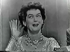 TV gif. In grainy black and white, a woman wearing a sparkling dress and ornate necklace rolls her eyes and gives a dismissive wave with a smile.