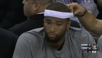 Sports gif. Demarcus Cousins sits on the bench next to teammates, looking doubtful and raising his fist half-heartedly.