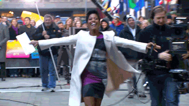 Video gif. Woman runs in heels with her arms spread out wide. She smiles happily and holds a paper in her hand. There’s a camera man behind her running with her and a crowd of people behind a barrier watching.