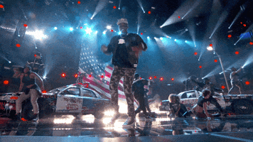 Video gif. Man performs at the BET Awards 2015. On stage there are graffitied cars and a large American flag. Everyone on stage falls to the ground like they’re dead.