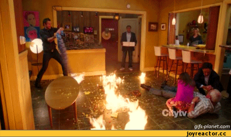 Video gif. Chaotic indoor scene where a man spins a fiery swath of fabric in a circle in front of him while other people appear stunned, and two people attend to a man appearing to be having a medical emergency on the floor, which is also on fire.