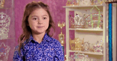 Reality TV gif. Kailia Posey on Toddlers and Tiaras sits in a room full of sparkly, extravagant tiaras. She looks away from us with big eyes and a cheeky smile.