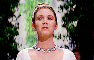 Star Wars gif. Carrie Fisher as Princess Leia stands regally with a large silver necklace and her braided hair wrapped into a bun. She can't help but grin shyly upon seeing someone.