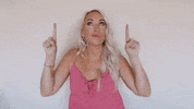 Video gif. Chelsie Kenyon, a marketing coach, pouts her lips and looks upwards while also pointing upwards, giving a little bouncy dance to emphasize her pointing. 