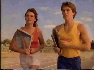 Video gif. Girl and boy smile at us as they run along a sunny track in tank tops and shorts while opening books with colored pages.