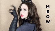 Celebrity gif. Lillee Jean dressed as Catwoman, faces us in profile, as she makes a claw with her hand and scratches down, staring at us with her teeth showing. Text, "Meow."