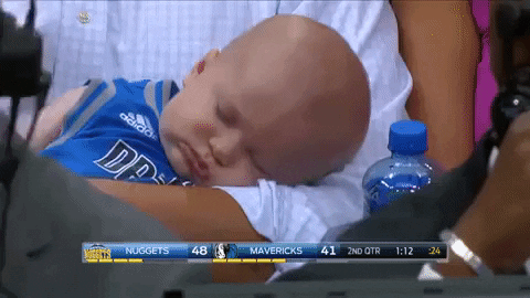 Sports gif. A baby wearing a jersey sleeps in the crook of an arm as a sportscaster works in the foreground and a score appears at the bottom of the screen.
