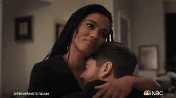 TV gif. Ryan Eggold as Dr. Dan and Freema Agyeman as Dr. Helen are hugging closely on a couch. He squishes closer to her and she rubs his back, with a soft smile on her face. 