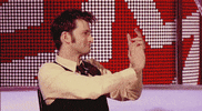 Celebrity gif. David Tennant sits and looks to his right, clapping.