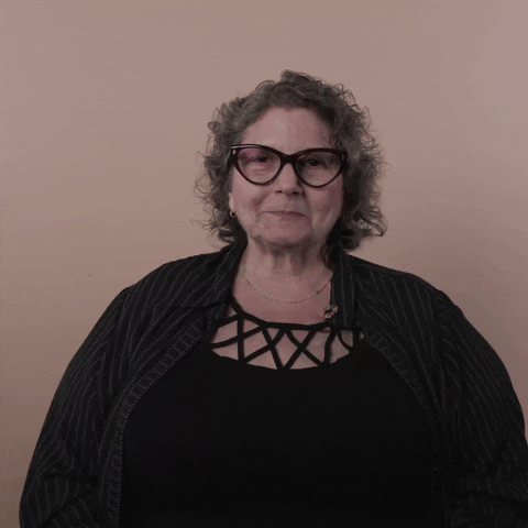 Reaction gif. A Disabled white woman with kinky curly gray hair and big wine-colored cat-eye glasses claps excitedly, mouth agape with glee.