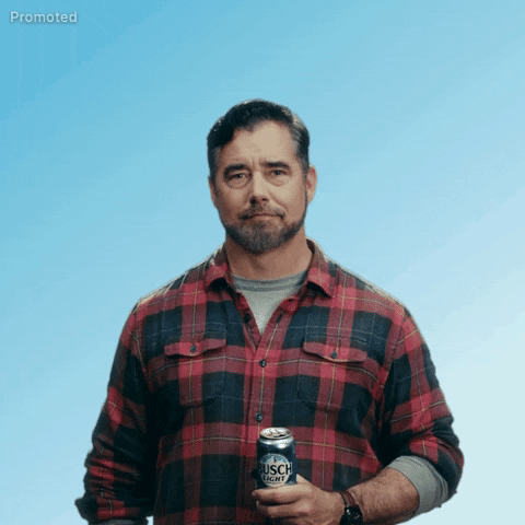 Sponsored gif. Gerald Downey wearing a red and black plaid shirt stands in front of a cloudless sky as he lifts a can of Busch Light beer to his chest, giving us a relaxed cheers with a cordial nod and unassuming side smile. Text, "Cheers."