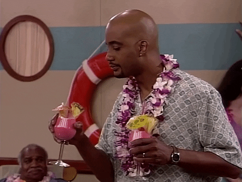 TV gif. John Henton as Overton in Living Single. He's on vacation and is on a cruise ship with a flower lei on. He's double fisting two cocktails and he sips on one slowly while raises his eyebrows in satisfaction at the taste.