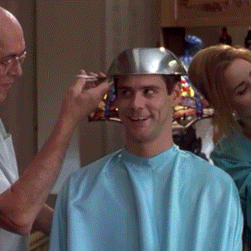 Movie gif. Jim Carrey as Lloyd from Dumb and Dumber looks around with a goofy smile while getting a haircut. The barber holds a metal bowl on Lloyd's head, and spins him around while trimming along the edge.