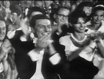 Celebrity gif. Black and white footage of Dick Van Dyke wearing a tuxedo as he smiles and claps in a crowd at an awards ceremony.