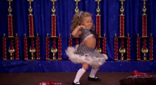 Video gif. A young girl in a blue checkered dress dances fiercely in front of a row of trophies. 