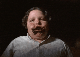 Movie gif. Jimmy Karz as Bruce Bogtrotter in Matilda sits on a chair and stares at us in a daze while chewing a mouthful of chocolate cake. He has chocolate frosting smeared all over his lips.