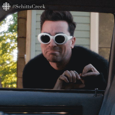 Schitt's Creek gif. Dan Levy as David wearing white-rimmed sunglasses with a fuzzy black sweater is overwhelmed with excitement as he stoops to speak us through the car window. Text, Can't wait.
