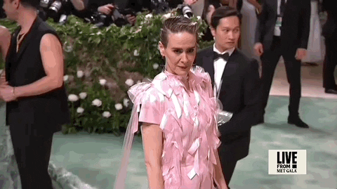 Met Gala 2024 gif. Camera zooms out on Sarah Paulson's pale pink short-sleeved Prada gown with a straight silhouette and long, thin panels of tulle that extend from her shoulders to the ground. The dress is covered in columns of pale pink satin bows that catch the light.