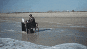 Video gif. Man works at a desk on the shore of a beach, continuing to type even as he stands up when the tide rolls in.