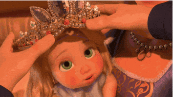 Disney gif. Baby Rapunzel from Tangled has an oversized tiara placed on her head and giggles.