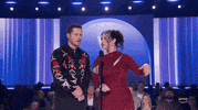 Academy of Country Music Awards gif. Noah Reid and Ashley McBryde share a standing microphone. Noah uses one arm to hold the other, in an unsure manner, while nodding head and seemingly saying "ahhh'. Simultaneously McBryde reaches out arm as she receives a ukelele from somone off camera while smiling at Reid as if fit was meant to surprise him.