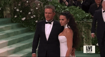 Met Gala 2024 gif. Matt Damon and his wife Luciana Barroso pose for pictures with Damon resting one hand at her back. Damon is wearing a black tuxedo, white button down shirt, and black bowtie. Barroso is wearing a white gown with a sweetheart neckline and a white collar that sweeps up and around her neck from the back of her dress. 