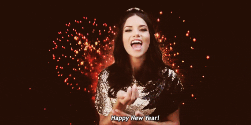 Video gif. As fireworks go off in the background, a woman in a sequined shirt holds up a handful of confetti and blows it at us. Text, "Happy New Year!"