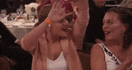 Video gif. At the Streamy Awards a young woman looks over her shoulder and raises her hands in a wave as people around her clap. 