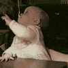 Video gif. A baby is holding a spoon in its mouth and they're trying to reach for it but failing, not understanding how the spoon is staying upright in its mouth. They've just discovered their lip muscles.