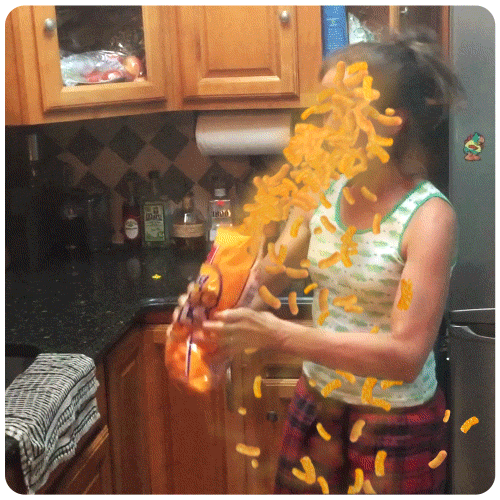 Video gif. A woman flinches as she holds a bag of cheese puffs that seems to be shooting out of the bag in a stream hitting her face. 