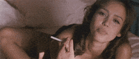 Celebrity gif. Holding a cigarette, Jessica Alba seductively rolls onto her belly in bed and smiles at us.