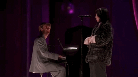 Oscars 2024 GIF. Billie Eilish and Finneas performing "What Was I Made For" on stage at the Oscars. They finish the song and Eilish holds her hands together in front of her and gives a slight bow of appreciation at the standing ovation. Finneas looks at her with pride and claps strongly for her as well.