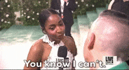 Met Gala 2024 gif. Closeup of Ayo Dibiri in an interview smiling and shaking her head as she responds to a question with the subtitle that appears in frame, "You know I can't." She's wearing a white halter backless Loewe gown covered in a textured lace pattern.