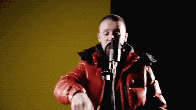 Video gif. A UK rapper on Daily Duppy is wearing a red puffer jacket and he tosses an imaginary ball with his right hand to his left which transforms into the word, "Yea."