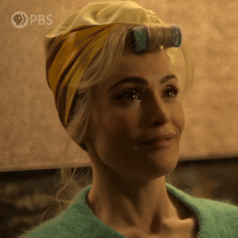 TV gif. Gemma Arterton as Barbara Parker on Funny Woman. She's wearing a yellow head wrap and a curler. She has a green sweater on and she splutters out laughing, covering her mouth at the last minute. 