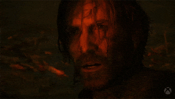 Video game gif. Close-up of a man with long disheveled hair and a beard in the game "Alan Wake." He mumbles or chants softly, looking off towards a red glowing light that illuminates his face. 