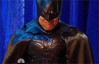 Video gif. Man in a Batman costume scrunches his face in a frown as he bows his head and squeezes the bridge of his nose.