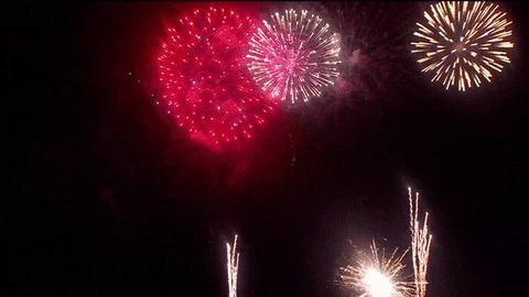 Video gif. Fireworks shoot up into a black sky, bursting into colors of red, orange, purple, and green. 